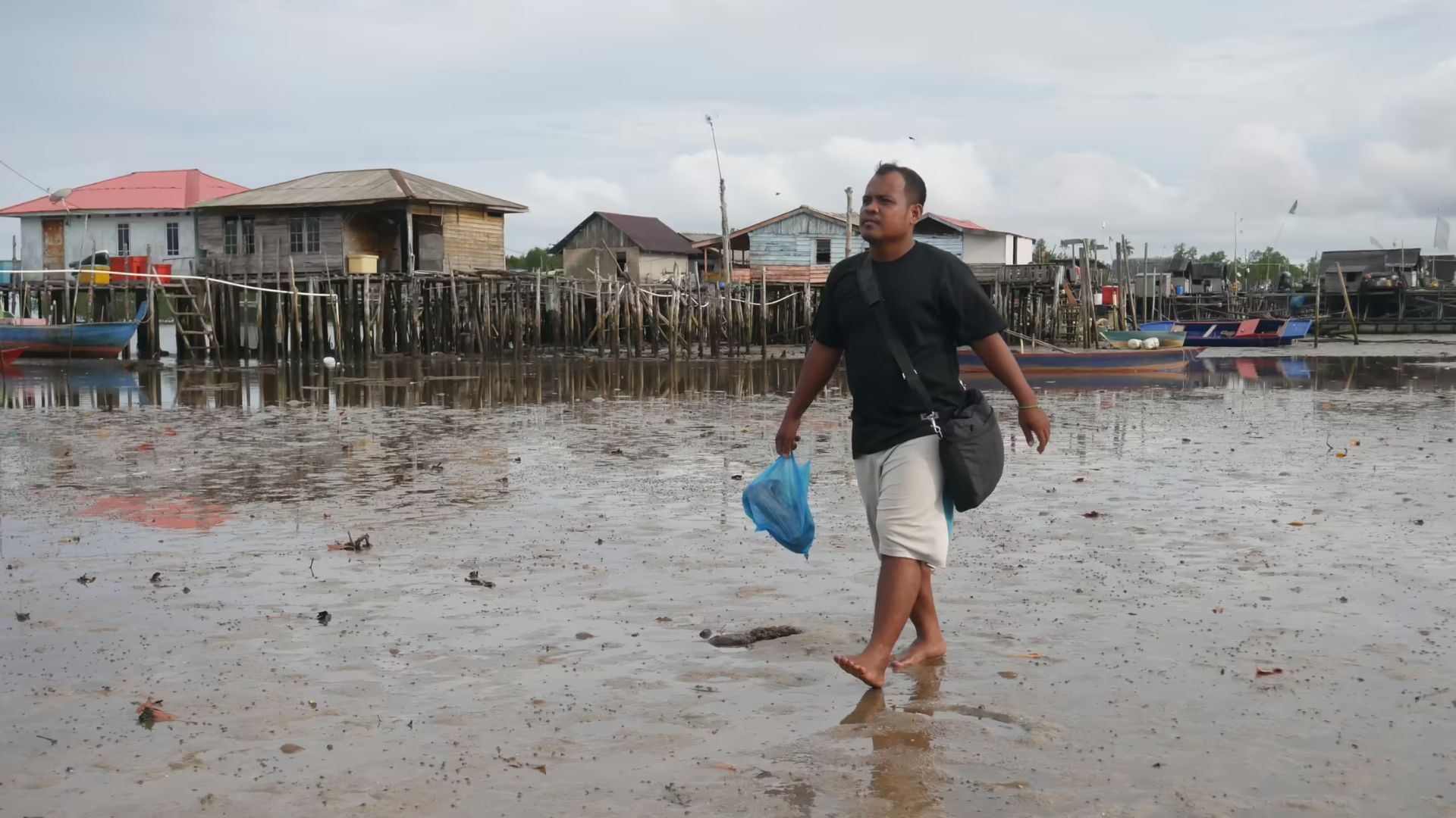 Johanes Jamil melintasi kawasan pesisir saat air laut surut dengan latar belakang pemukiman Suku Laut, Kawal Laut, Bintan, Kepulauan Riau, Sabtu (15/3/2025). Kawasan ini menjadi tempat tinggal bagi komunitas Suku Laut yang masih bergantung pada laut sebagai sumber penghidupan utama.